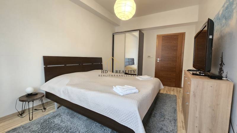 HD REALITY, Vám ponúka na Predaj 3-izbový Apartmán + Garážové státie - Spálňa v 3-izbovom byte s manželskou posteľou, televízorom a podlahou s dreveným dekorom.