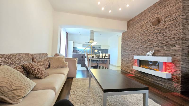 HD REALITY, Vám ponúka na Predaj 3-izbový Apartmán + Garážové státie - Obývačka v 3-izbovom byte s podlahou s dreveným dekorom, dekoratívnou stenou a krbom.