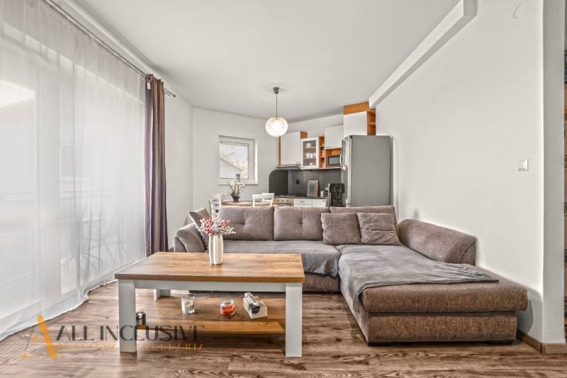 NA PREDAJ 2-IZBOVÝ BYT S 2 BALKÓNMI, DUNAJSKÁ STREDA - Obývačka v 2-izbovom byte so sedačkou, kuchynskou linkou a podlahou s dreveným dekorom.