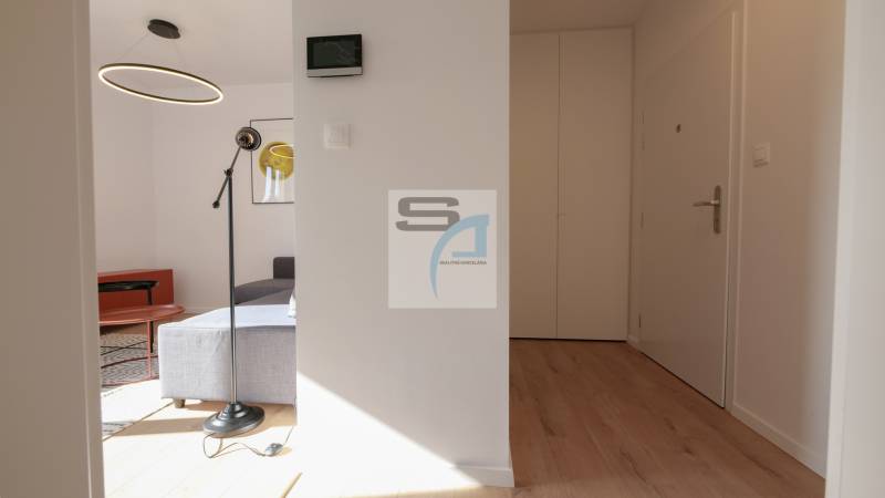 2-izbový byt s terasou 15 m² a parkovaním v tichej časti Bratislavy - Interiér 2-izbového bytu s podlahou s dreveným dekorom, kruhovým lustrom a sivou pohovkou.