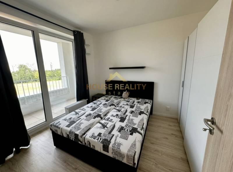 Nový 2 izbový byt SLNEČNICE, Labutia ulica, Bratislava V. Petržalka - Spálňa v 2-izbovom byte s posteľou, skriňou a balkónom s podlahou s dreveným dekorom.