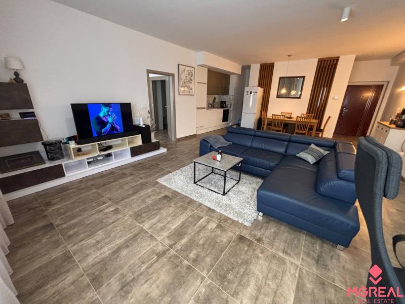 Veľký 96 m2  2 izbový byt s garážovým státím Topoľčany Centrum  - Obývacia izba v 2-izbovom byte s tmavou sedačkou a televízorom, podlaha s dreveným dekorom.