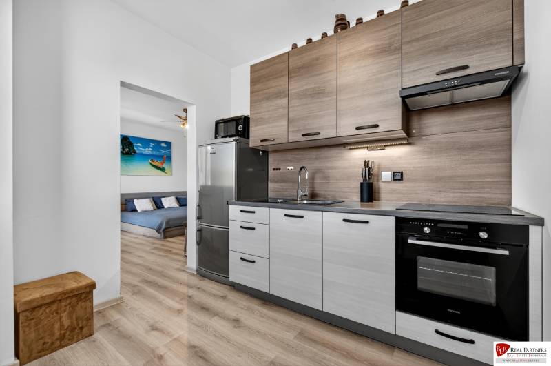REZERVOVANÉ Predaj moderný 1-izbový byt, 45,24m2, Bratislava IV - Kuchyňa v 1-izbovom byte s chladničkou, dekorom dreva a obrazom pláže v pozadí.