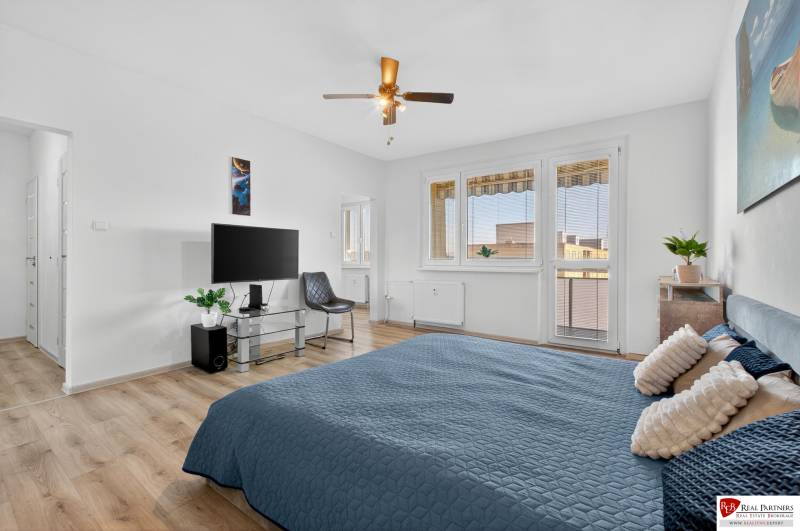 REZERVOVANÉ Predaj moderný 1-izbový byt, 45,24m2, Bratislava IV - Spálňa s posteľou, televízorom, kreslom, rastlinou a podlahou s dreveným dekorom v 1-izbovom byte.