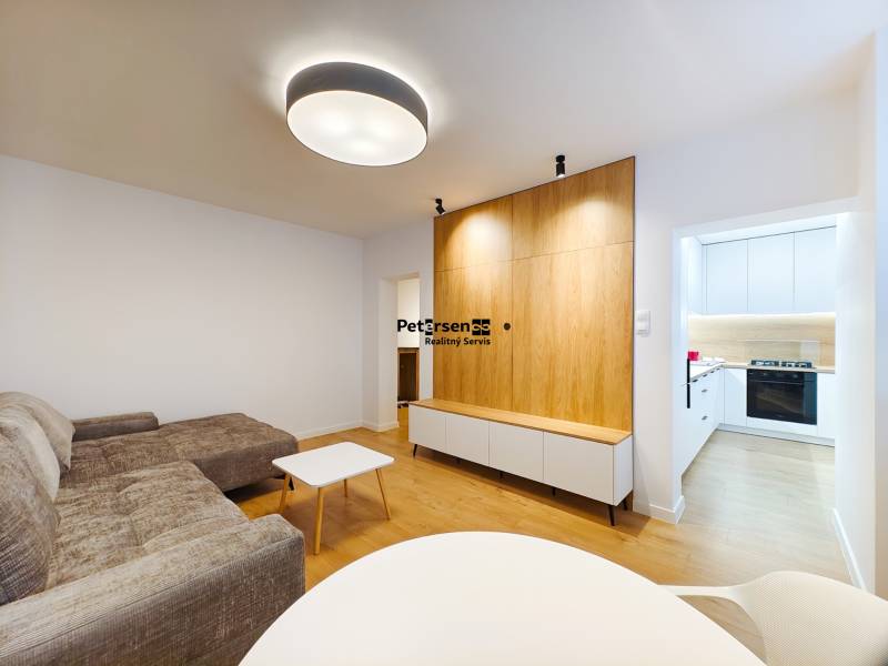 Prenájom úplne nového 2 izb. bytu - skvelá poloha a komfortné bývanie. - Obývacia izba v 2-izbovom byte s podlahou s dreveným dekorom, vstupom do bielej kuchyne.