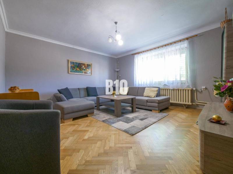 Rodinný dom s krásnym veľkým pozemkom ( stavebným) - Nové Sady - Obývacia izba v rodinnom dome s hnedou podlahou, šedou sedačkou a veľkým oknom.