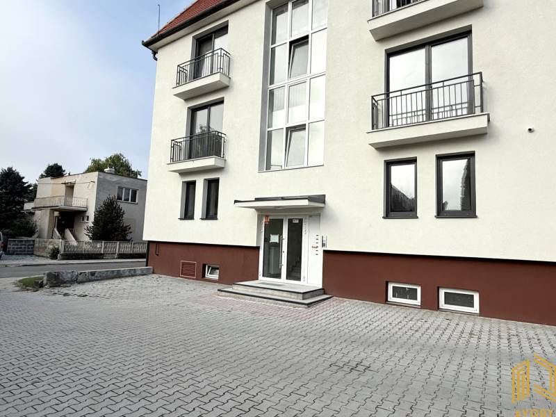 NOVÝ 2 - IZB. BYT (63m2), SENICA - SADOVÁ - Budova na Sadovej ulici v Senici s bielou fasádou a dlaždeným parkoviskom.