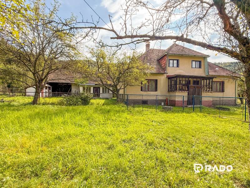 5i RD s garážou, Čierna Lehota, okr. Bánovce n./B. - Rodinný dom v Čiernej Lehote s trávnikom, stromami a oplotením na pozemku.