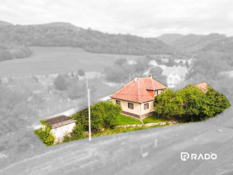 5i RD s garážou, Čierna Lehota, okr. Bánovce n./B. - Rodinný dom v Čiernej Lehote obklopený zeleňou, mierne kopcovitá krajina v pozadí.