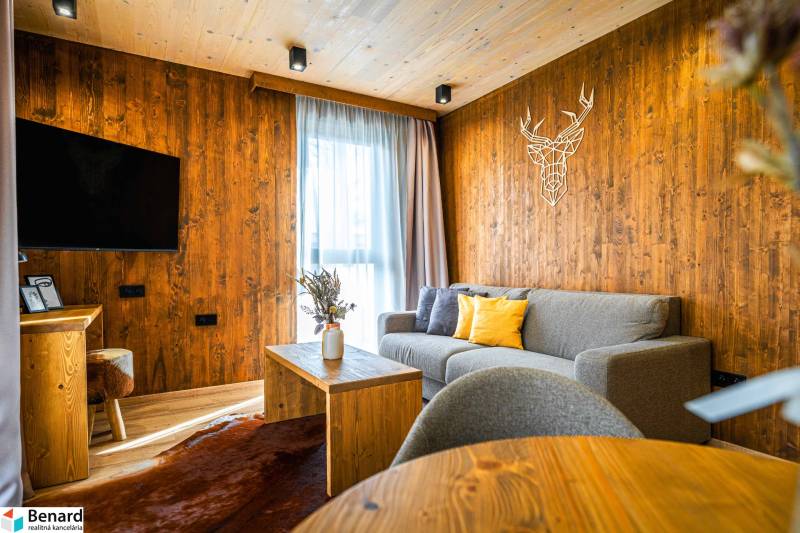 Na predaj krásny apartmám v Štrbe - Obývacia izba s drevenými stenami, sivou pohovkou a podlahou s dreveným dekorom v 2-izbovom byte.