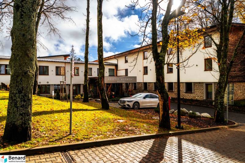 Na predaj krásny apartmám v Štrbe - Budova hotela v Štrbe na Lúčnej obklopená stromami, so zaparkovaným autom.