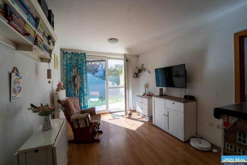 Na predaj útulný 2 izbový byt so zahradkou Svätý Jur 165.000,- Eur - Obývacia izba v 2-izbovom byte s podlahou s dreveným dekorom, televízorom a kreslom.