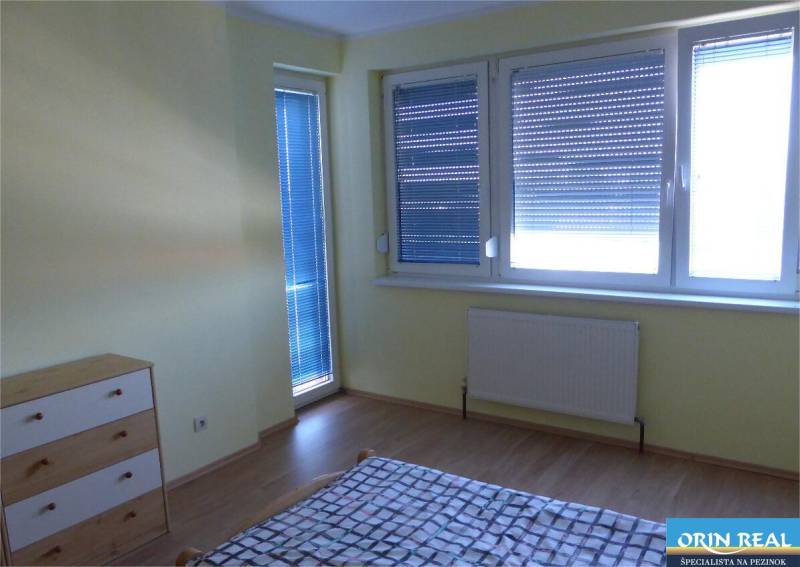 Na predaj 2 izbový byt s parkovacím státím Zemplínska ul. - Izba v 2-izbovom byte s podlahou s dreveným dekorom, žalúziami a nábytkom.