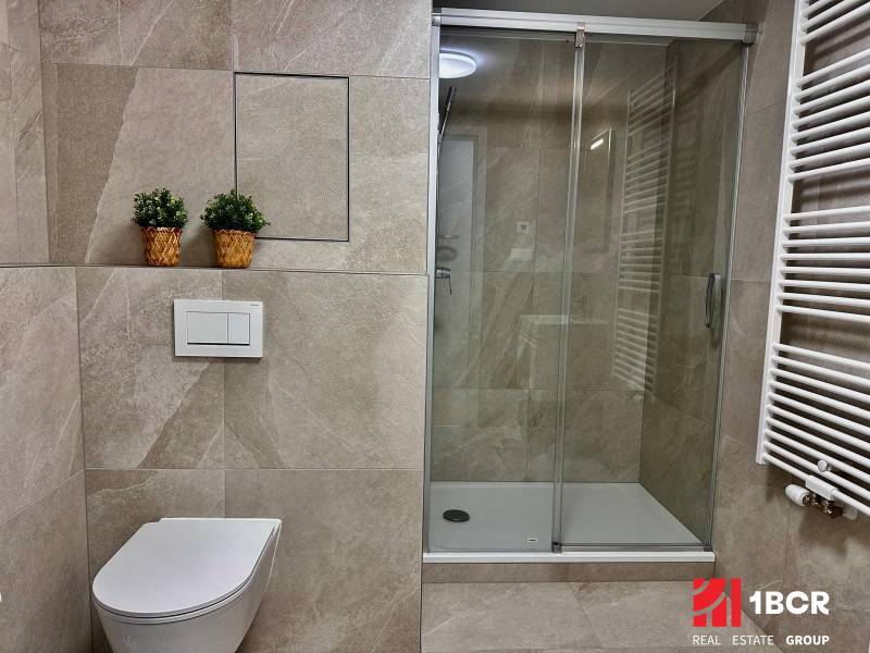 PRIESTRANNÝ 1-IZB. BYT S LOGGIOU V IKONICKOM PROJEKTE ZWIRN III - Kúpeľňa v 1-izbovom byte s toaletou, sprchovým kútom a živými rastlinami v košíkoch.