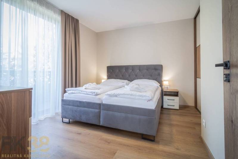 Lukratívne dvoj a štvorizbové byty v Tatranskej Lomnici na prenájom - Spálňa v 2-izbovom byte s pohodlnou posteľou, podlahou s dreveným dekorom a závesmi.