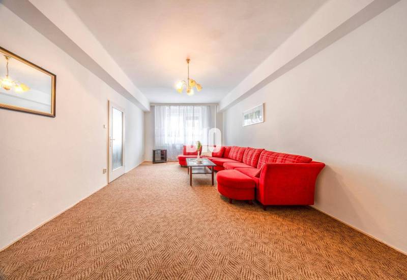 KRÍŽNA: VEĽKORYSÝ 4-IZBOVÝ BYT S RETRO ŠARMOM BLÍZKO CENTRA - Obývacia izba so svetlou stenou, červenou sedačkou a kobercom v 4-izbovom byte.