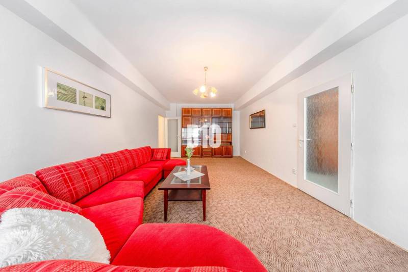 KRÍŽNA: VEĽKORYSÝ 4-IZBOVÝ BYT S RETRO ŠARMOM BLÍZKO CENTRA - Červená pohovka, sklenený stolík, vstavaná skrinka a svetlý koberec v 4-izbovom byte.