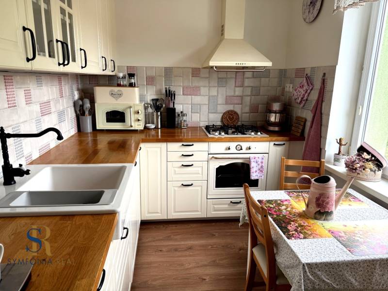 2-izbový byt v Seredi- bez rezervačnej zálohy - Kuchyňa 2-izbového bytu s podlahou s dreveným dekorom, biele skrinky, kvety a dekorácie.