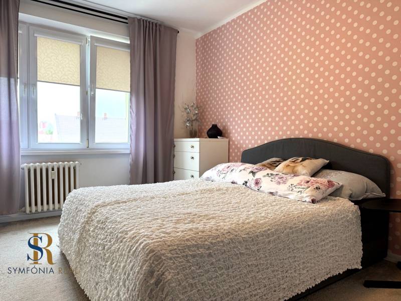 2-izbový byt v Seredi- bez rezervačnej zálohy - Spálňa s bodkovanou tapetou v 2-izbovom byte s veľkými oknami a kvetinovými vankúšmi.