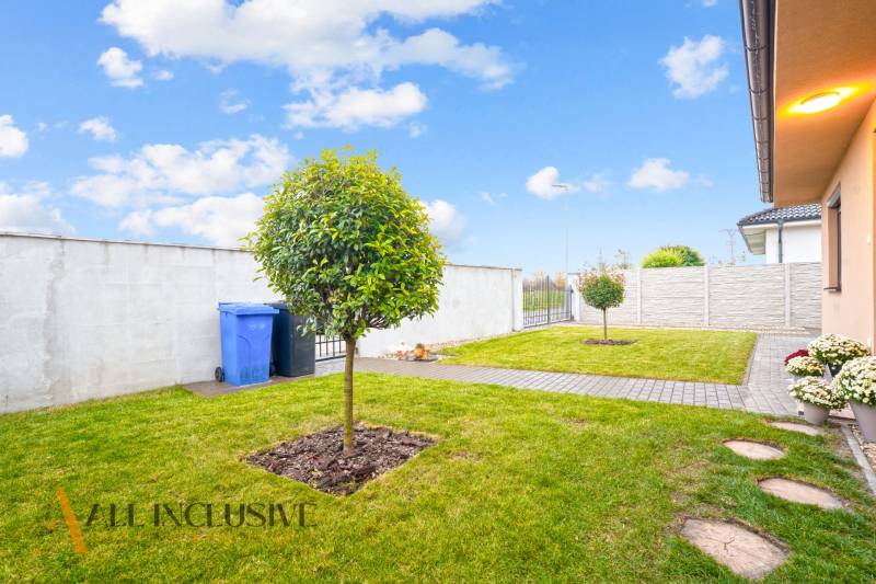 NA PREDAJ MODERNÝ 3-IZB. RD TYPU BUNGALOV V GABČÍKOVE - Záhrada rodinného domu v Gabčíkove s trávnikom, stromom a chodníkom vedľa plotu.