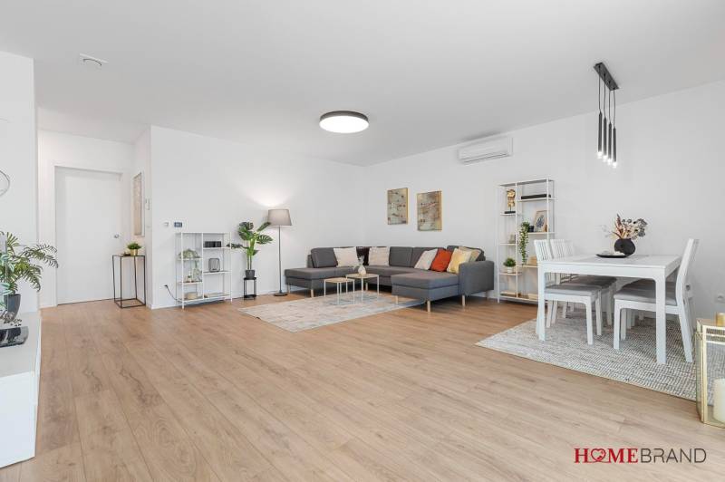 BÝVANIE VO VILA DOME - POSLEDNÝ VOĽNÝ 3-IZBOVÝ BYT SO ZÁHRADOU - Priestranná obývacia izba s podlahou s dreveným dekorom a sedačkou v 3-izbovom byte.