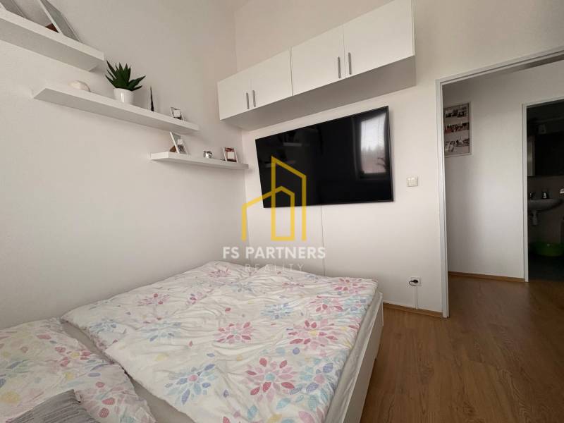 Prenájom moderného 2-izbového bytu v centre Komárna, 40 m² – Petőfiho  - Svetlá spálňa v 2-izbovom byte s TV a podlahou s dreveným dekorom.