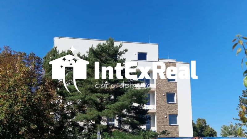 NOVOSTAVBA -2 izbový byt s komorou, na predaj  Galanta - Budova na Športovej ulici v Galante, obklopená stromami, s jasnou oblohou v pozadí.