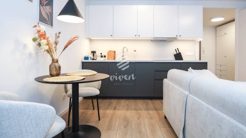 PRENÁJOM - 1 izbový byt v projekte Ovocné sady - Kuchyňa v 1-izbovom byte s bielymi skrinkami, tmavou linkou a podlahou s dreveným dekorom.