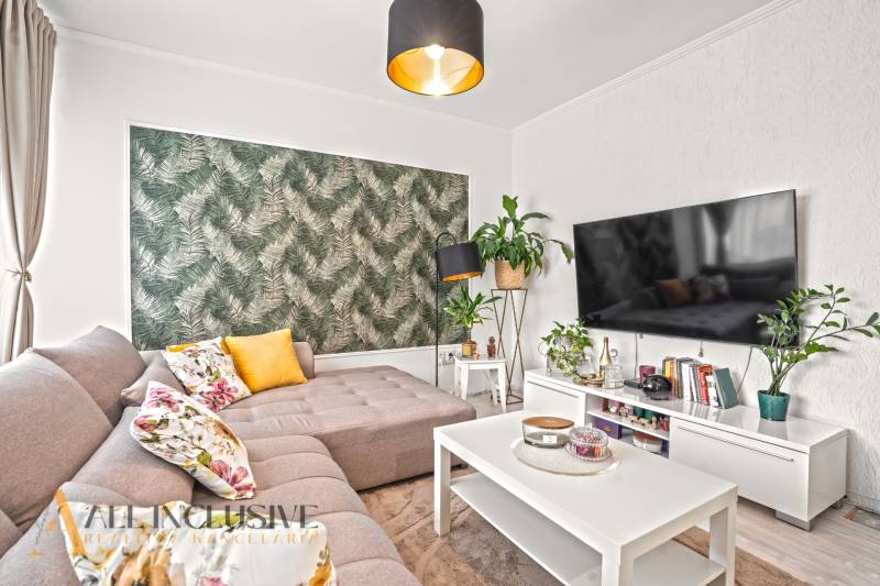 NA PREDAJ 3-IZB. BYT S TERASOU A ZÁHRADKOU, DUN.STREDA - Svetlá obývačka s pohovkou a kvetinovými vankúšmi, zelená tapeta, televízor na stene.