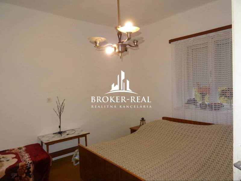 Rodinný dom, chalupa 2 Izbový - Fony - Spálňa v rodinnom dome s posteľou, stolíkom a lustrom, na stene je okno so záclonou.