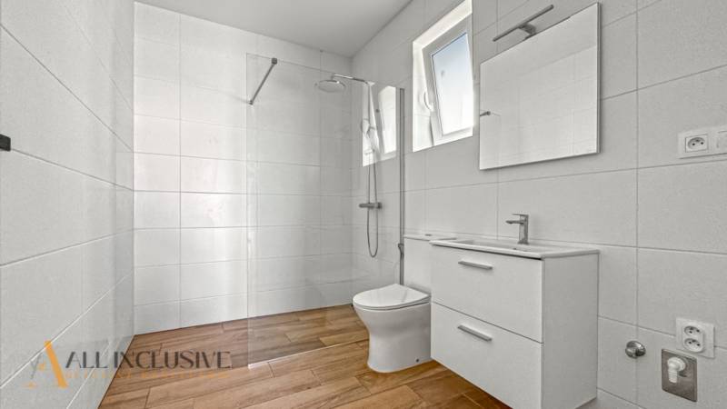 IDEÁLNY 1 IZBOVÝ BYT | VEĽKÉ RIPŇANY | TOPOĽČANY - Kúpeľňa s bielymi obkladačkami, sprchou a podlahou s dreveným dekorom v 1-izbovom byte.