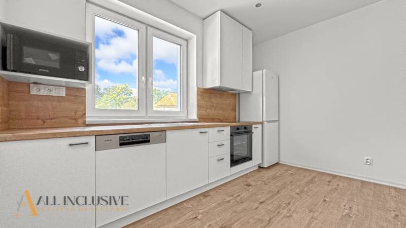 IDEÁLNY 1 IZBOVÝ BYT | VEĽKÉ RIPŇANY | TOPOĽČANY - Svetlá kuchyňa s bielymi skrinkami, podlaha s dreveným dekorom, okno s výhľadom.