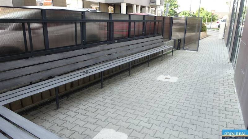 PRENÁJOM NEB.PRIESTORU V POLYFUNKČNOM DOME V CENTRE PEZINKA - Dlhá lavička na Moyzesovej ulici pred obchodnými priestormi s dlažbou a zábradlím.