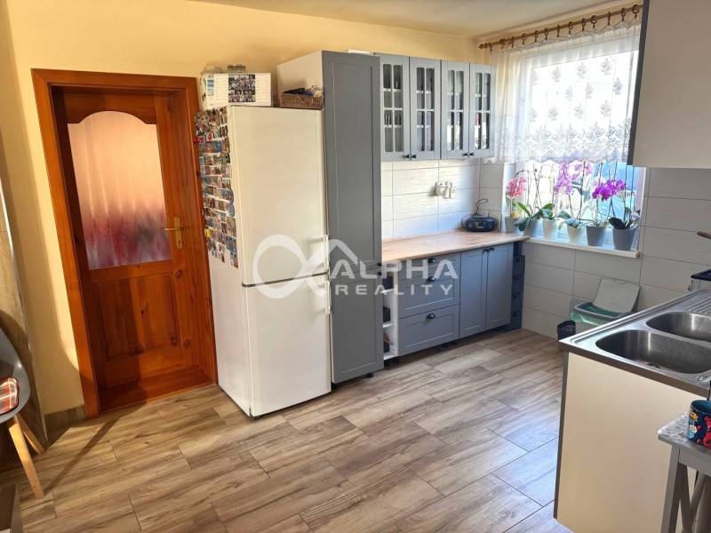 EXKLUZÍVNE Rodinný dom, širšie centrum, Spišská Nová Ves - Kuchyňa v rodinnom dome s bielymi spotrebičmi, kvetmi na parapete a podlahou s dreveným dekorom.