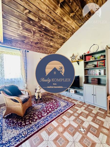 Vám ponúka na predaj rodinný dom v Komárne, ča - Interiér rodinného domu s dreveným dekorom, koberec, kreslo a úložná skrinka.