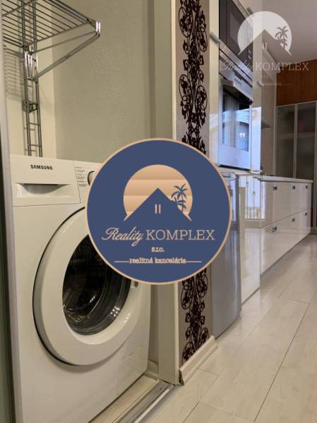 Vám EXKLUZÍVNE ponúka NA PRENÁJOM moderný 2 iz - Kuchyňa v 2-izbovom byte s práčkou, bielym nábytkom a podlahou s dreveným dekorom.