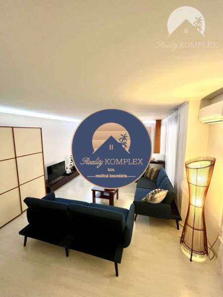 Vám EXKLUZÍVNE ponúka NA PRENÁJOM moderný 2 iz - Obývačka v 2-izbovom byte s podlahou s dreveným dekorom, modrými sedačkami a televízorom.