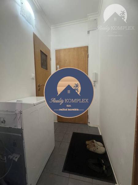 Vám ponúka 1 izbový byt vo VYHĽADÁVANEJ lokali - Vstupná chodba v 1-izbovom byte s práčkou a obuvou na tmavej dlažbe.