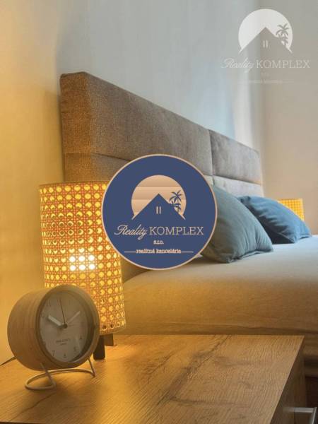 Vám ponúka 1 izbový byt vo VYHĽADÁVANEJ lokali - Čiastočný pohľad na posteľ v 1-izbovom byte s lampou a budíkom.