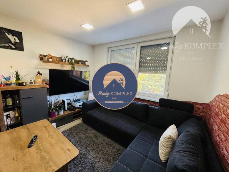 Vám ponúka exkluzívne na predaj moderný, komple - Obývacia izba s tmavou sedačkou, televízorom na stene a veľkým oknom.