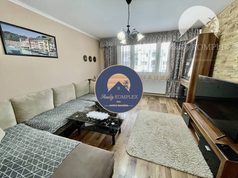 Vám ponúka 3-izbový byt s loggiou po kompletnej - Obývacia izba v 3-izbovom byte, so sedačkou, koberec, televízorom a podlahou s dreveným dekorom.