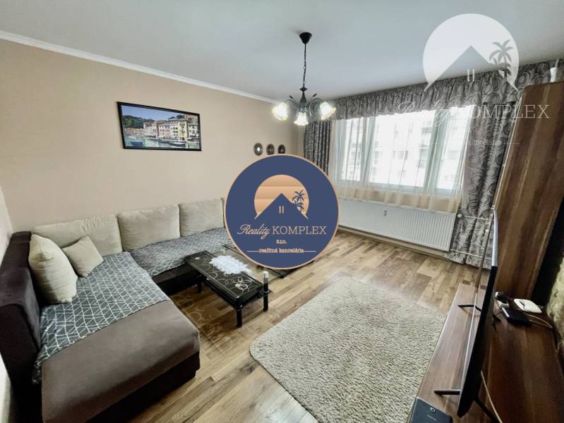 Vám ponúka 3-izbový byt s loggiou po kompletnej - Obývačka 3-izbového bytu so sedačkou a podlahou s dreveným dekorom.