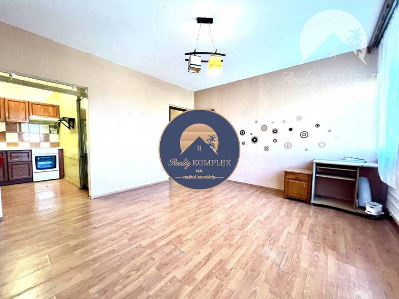 Ponúka na PREDAJ 1 izbový byt v Komárne - Izba s podlahou s dreveným dekorom, kuchyňou a jednoduchým nábytkom v 1-izbovom byte.