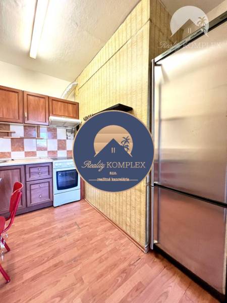 Ponúka na PREDAJ 1 izbový byt v Komárne - Kuchyňa 1-izbového bytu s podlahou s dreveným dekorom a nerezovou chladničkou.