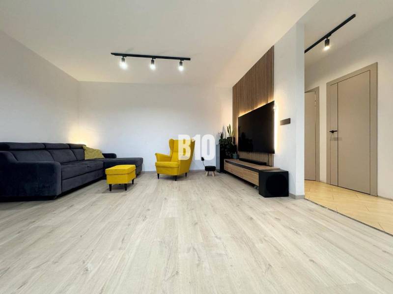 Veľkometrážny 3 izbový byt s garážou - Obývačka v 3-izbovom byte s podlahou s dreveným dekorom, žlté kreslo, rohová sedačka.