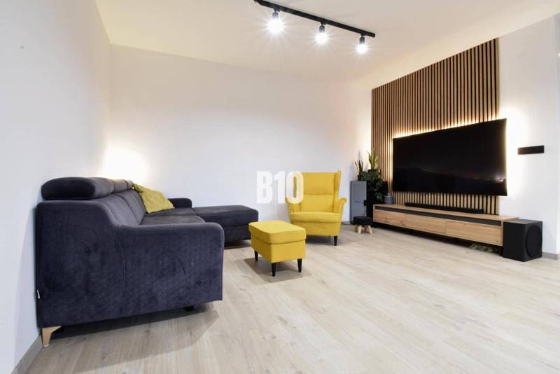 Veľkometrážny 3 izbový byt s garážou - Obývacia izba v 3-izbovom byte s podlahou s dreveným dekorom, TV a nábytkom.