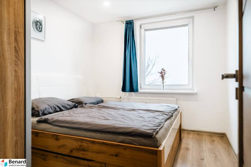 Útulný 2-izbový byt na prenájom - Spálňa s podlahou s dreveným dekorom a posteľou v 2-izbovom byte, modrý záves na okne.