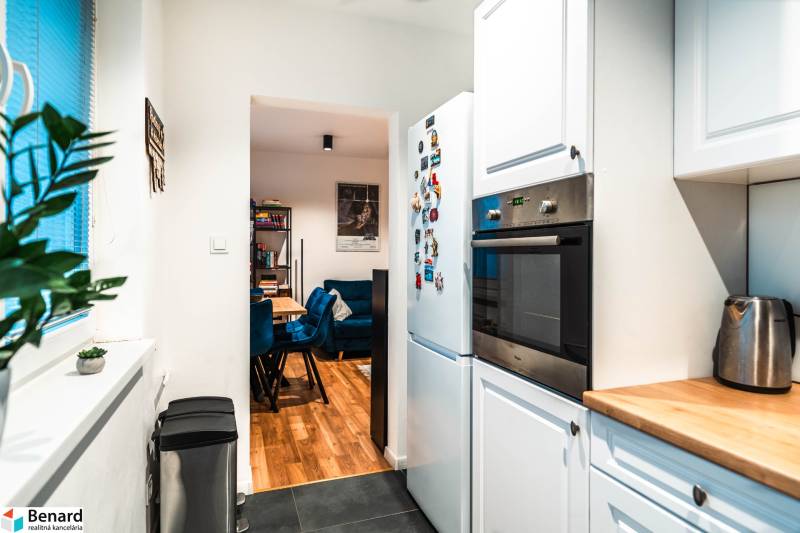 Na prenájom moderný 2-izbový byt  - Kuchyňa 2-izbového bytu s bielymi skrinkami, spotrebičmi a výhľadom do jedálne.