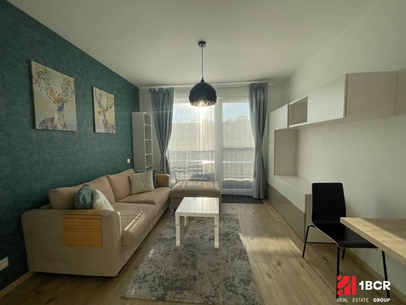 MODERNÝ, KOMPLETNE ZARIADENÝ 1-IZBOVÝ BYT V PROJEKTE SLNEČNICE - Obývačka 1-izbového bytu s pohovkou, dekoráciou jeleňa a podlahou s dreveným dekorom.