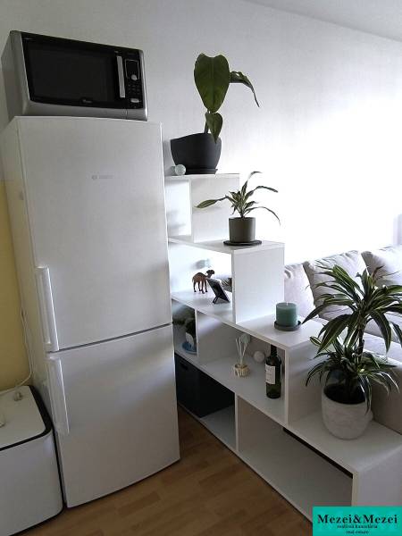 Na dlhodobý prenájom 2-izbový byt - kompletne zariadený, klimatizovaný - Kuchynský kút v 2-izbovom byte s bielou chladničkou a rastlinami na poličkách.